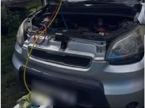 Recharge de clim en cours, tuyaux branchés sous le capot d'une voiture garée sur l'herbe